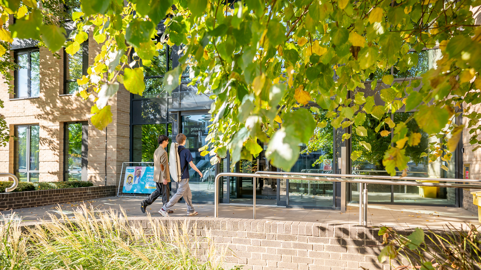 Two men walking towards an entrance on a sunny day with a green tree and grass.